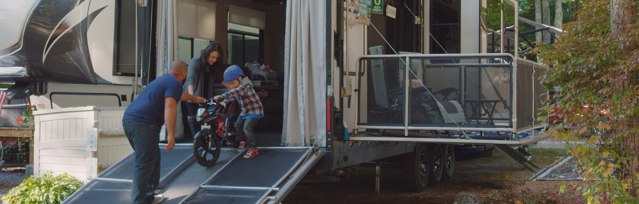 Parents helping a young kid walk a bicycle down a toy hauler ramp.